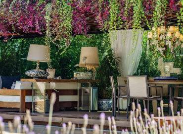 Pergolado envolto por plantas, com mesa, sofá e abajours.