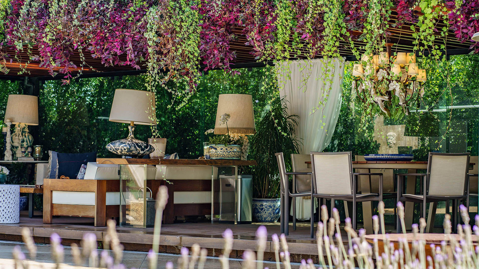 Pergolado envolto por plantas, com mesa, sofá e abajours.