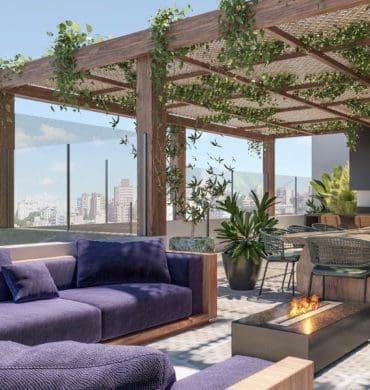 Rooftop com vista para cidade, pergolado com folhagens, lareira e sofá estofado na cor azul, mesa de jantar retangular na cor marrom claro e cadeiras na cor verde.