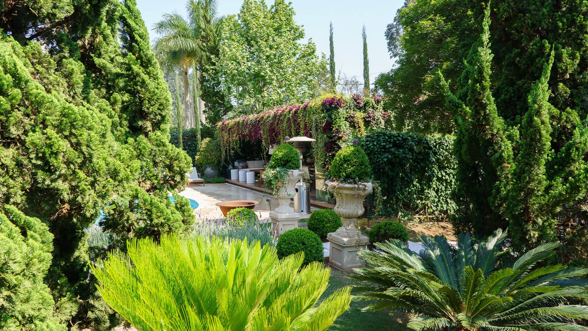 A foto mostra um exuberante jardim com ciprestes, arbustos e ao fundo um pergolado coberto por plantas.