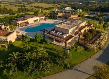 Vista superior de condomínio de alto padrão, com clube, piscina e mais de 200 mil m² de natureza. Local com paisagismo mediterrâneo apresenta diversas espécies, como tamareiras-das-Canárias, cicas, capins-do-Texas e juníperos-rasteiros.