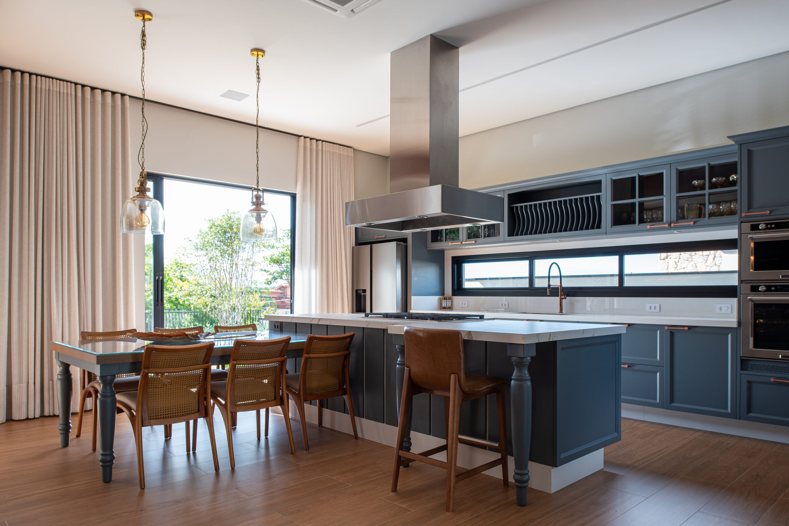 Cozinha com armátios em laca azul petróleo e bancada dekton, piso de madeira e ao fundo uma porta com área verde e cortinas sob ela a frente mesa de jantar com cadeiras e bancada de madeira