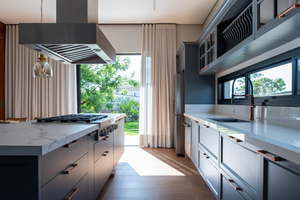 Cozinha com armátios em laca azul petróleo e bancada dekton, piso de madeira e ao fundo uma porta com área verde e cortinas sob ela