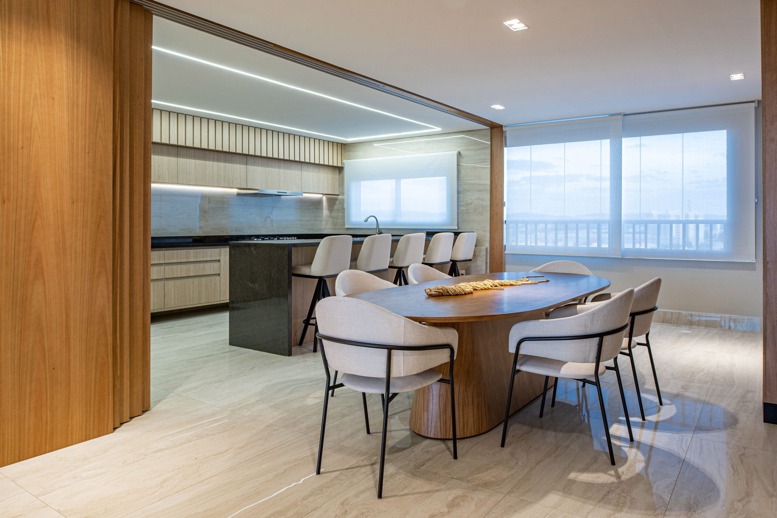 Sala de jantar com mesa de madeira e cadeiras modernas com estofado off white, interligada a cozinha com bancada em mármore escuro, com banquetas na cor off white.