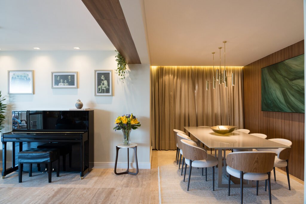 Sala de jantar com mesa grande ao lado de um quadro verde em uma parede de madeira, piano preto ao lado e uma mesa lateral com flores amarelas